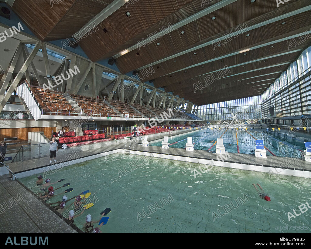 SIMON INGLIS. Crystal Palace National Sports Centre, Crystal Palace Park, Bromley, Greater London Authority, 2013. The swimming pool in the Crystal Palace National Sports Centre, with a lesson taking place in the learner pool in the foreground. The National Recreation Centre was opened in 1964. It was built on the site of an exhibitions centre of the Crystal Palace Trust. When the designs were drawn up in 1953-1954, it was the first indoor multi-functional hall in Britain, incorporating both wet and dry sports in one building. The swimming pool side of the building houses a 50m pool, diving pool and learner pool. The building is Grade II* Listed.
