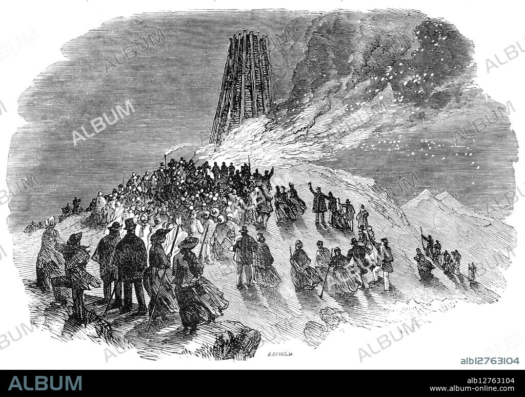 EDMUND EVANS. The lighting of the Malvern Monster Beacon, 1856. Illustration of a sketch by Cuthbert Bede. '...it was intended to have been a simple bonfire to commemorate the lighting of Malvern with gas...But the project increased in importance as it became more widely known...The Beacon...towered up like a huge Druid idol. Twelve iron-banded poplar trees...that narrowed to its summit kept together the matériel - 450 faggots, 5 cords of wood, 12 tar-barrels, 2 tons of coals, 3 or 4 loads of hop poles, 2 loads of furze, 2 barrels of tar, and 1 of naphtha...The effect of [the] torchlight procession as it wound up the zigzag path was exceedingly picturesque...by the time that the Beacon was lighted, upwards of 1500 people had assembled on the summit of the hill...At a quarter before seven a blue light was burnt, succeeded by a red light...a magnificent, flight of fifty rockets rushed into the heavens, and burst into a galaxy of falling stars - the signal to distant spectators of the lighting of the Beacon...[In] the hot vicinity of the monster fire...red and orange flames...rushed out longitudinally into wreathed masses of velvety smoke, and drove a fiery rain of sparks far into the darkness...over all, was the deep blue sky studded with silvery stars'. From "Illustrated London News", 1856.