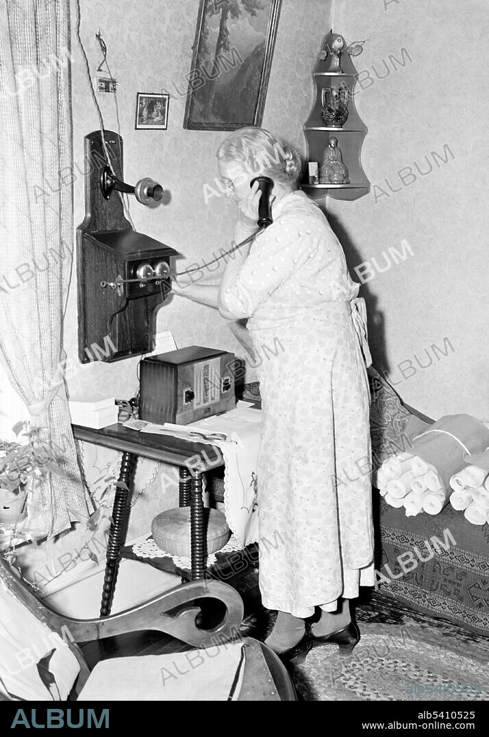 Lady signaling operator on old style telephone. One of the first telephones commercially available was the hand cranked wall telephone, which was basically a wooden rectangular box that housed the necessary components with a receiver that hung on the exterior. These wall telephones were referred to as camera telephones and box telephones. Photographed by John Vachon, 1940 (cropped and cleaned).