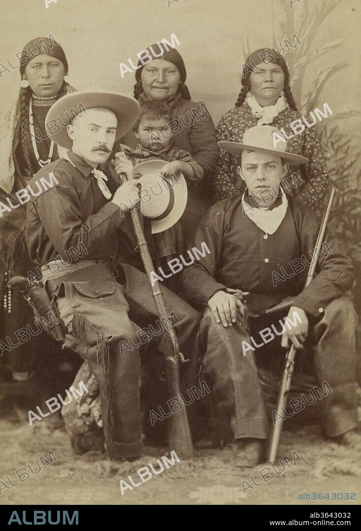 Two Fully Dressed Cowboys, Missoula, Montana Territory, Circa 1880S. Wild West Cowboys With Hats, Fringed Leather Chaps, And Cowboy Shirts With Kerchiefs. The Cowboy Seated On Left Has A Colt Revolver In A Double Loop Holster, As Well As A Percussion Rifle. The Very Young Cowboy On The Right Has What Appears To Be A Winchester In His Arms. Posed With Three Native American Women And A Child., James Ollason, 1880.