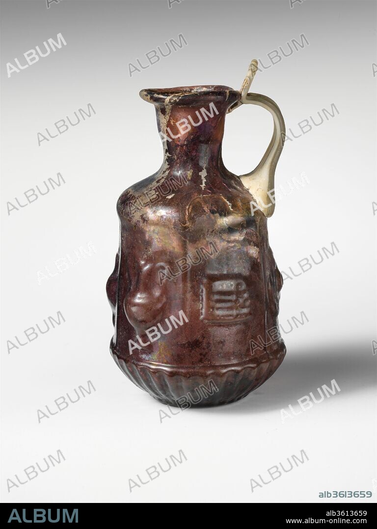 Glass hexagonal jug with Dionysiac symbols. Culture: Roman. Dimensions: H.: 3 7/8 in. (9.8 cm). Date: 1st century A.D..
Translucent deep purple, with handle in semi-opaque colorless with greenish tinge.
Tubular rim, folded out, round, and into flaring mouth; cylindrical neck, tapering downward; convex sloping shoulder; straight-sided hexagonal body, expanding downward; convex undercurve; flat circular bottom; strap handle attached in two large ribs to shoulder and top edge of body above panel with circular object, drawn up and out, then curved in, and pressed on to top of neck and underside of rim, with projecting flattened thumbrest above. Slight mis-alignment of vertical sections of mold on one side above cup section.
On shoulder, frieze of indistinct downturned tongues; on body, six rectangular panels, with vertical raised edges, containing objects associated with the Dionysiac cult: 1. crossed, double-ended thyrsi; 2. a footed jug with spout to left and high handle to right; 3. a footed amphora with high handles; 4. a syrinx (pan pipes) suspended horizontally from a vertical string; 5. a circular object with central protruding boss (a phiale mesomphalos or, perhaps, a cymbal); 6. a footed crater with high handles; around bottom, a frieze of thirty-one upturned, rounded tongues in raised relief; on bottom, raised circle around edge, a fine shallow circle surrounding a thicker and higher ring around a hollow central boss.
Broken, with several cracks and two holes, one in shoulder, the other in body below handle; handle streaked with black impurities; dulling, iridescence, and patches of whitish weathering on exterior, creamy brown weathering and iridescence on interior.