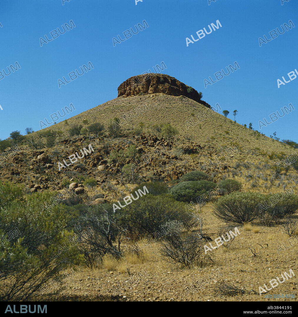 Desert mesa, a flat-topped hill with resistant cap rock surrounded by scree, is a typical desert landform in central Australia. The scree is vegetated by porcupine grass (Triodia sp.), green from a recent rain, and Cassia and Acacia shrubland pioneers on the lower slopes.