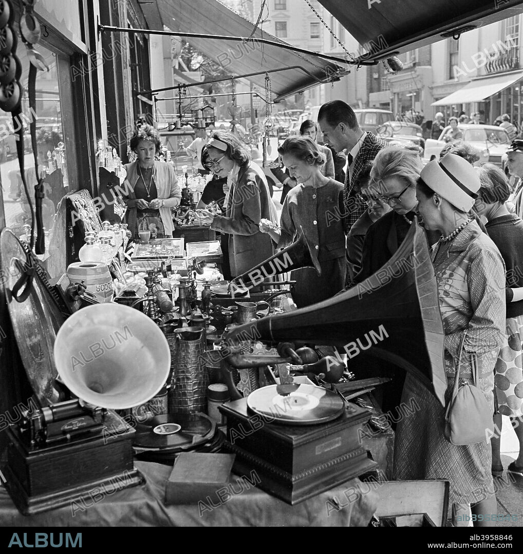 Street market, Portobello Road, London, 1962-1964. People crowded around a stall outside a shop selling gramophones and other items, while the stallholder looks on at the back.