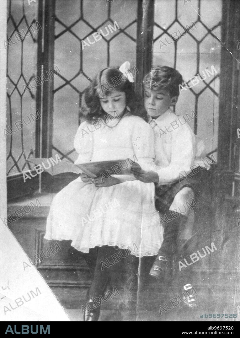 The Queen Mother as a young girl.. Reading a book with her brother, dressed for the drawing room.