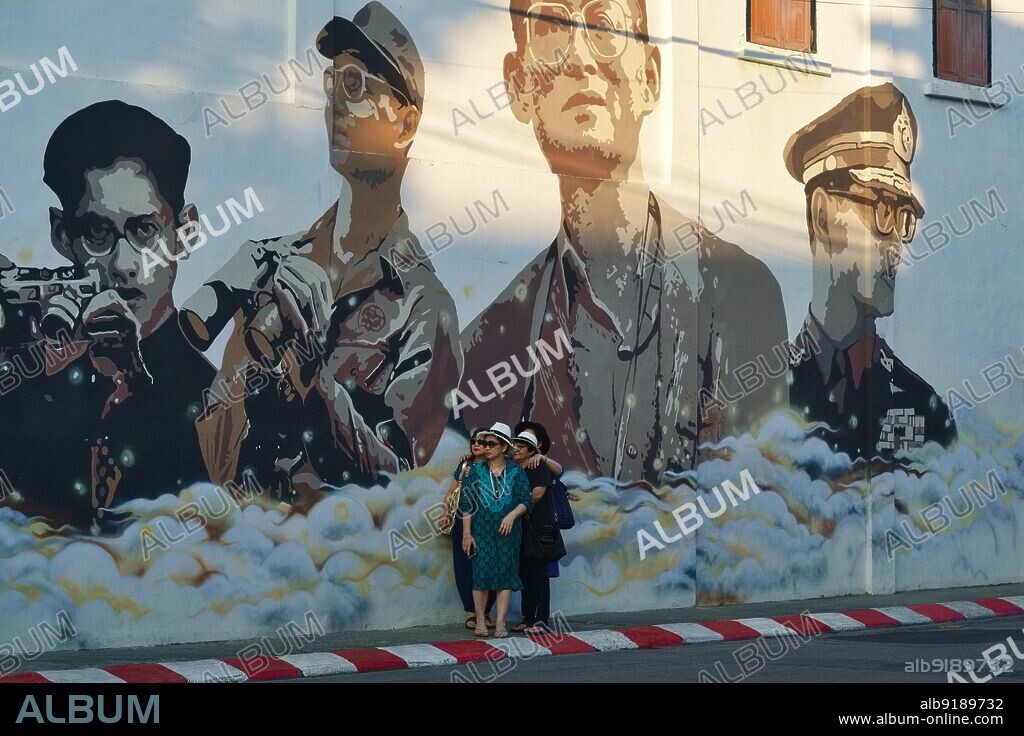 Thailand: Tourists in Phuket Town (Phuket City), posing under a wall painting depicting former Thai King Bhumibol Adulyadej (5 December 1927 Äì 13 October 2016), 9th monarch of the Chakri Dynasty, in different phases of his life. Bhumibol Adulyadej (Phumiphon Adunyadet) was the 9th King of Thailand. He was known as Rama IX, and within the Thai royal family and to close associates simply as Lek. Having reigned since 9 June 1946, he was one of the world's longest-serving heads of state and the longest-reigning monarch in Thai history.
