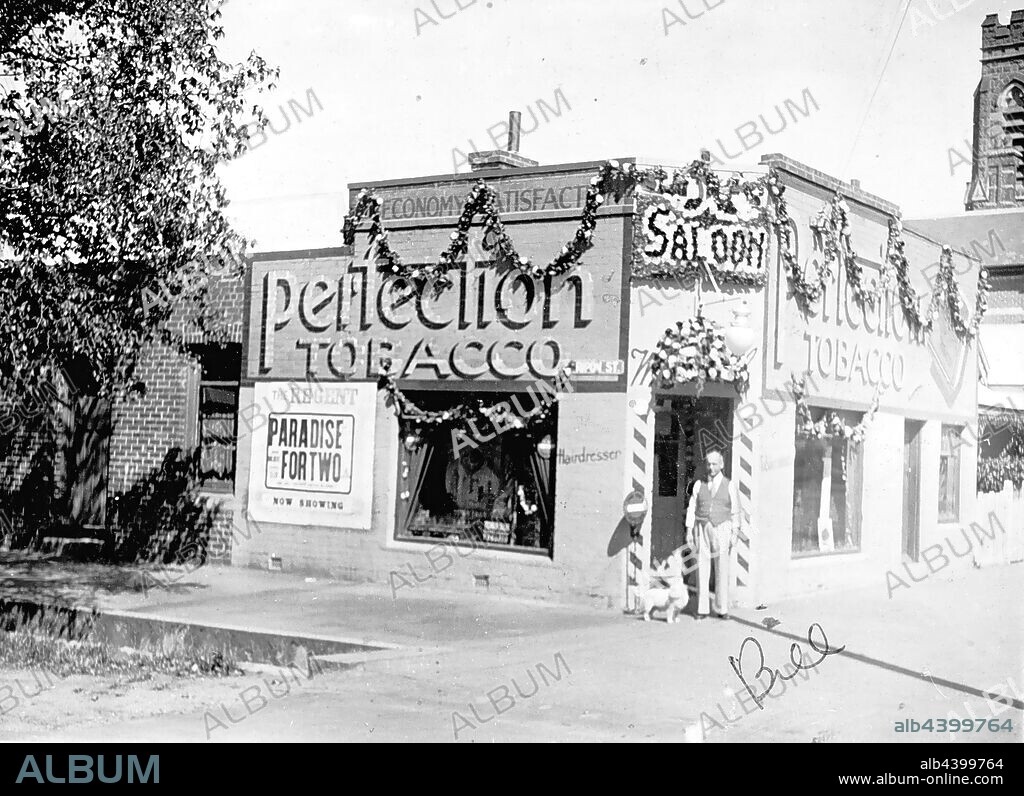 Negative - Ballarat, Victoria, 1938, A tobacconist's shop & barber's saloon decorated for the Ballarat centenary celebrations. St Peter's Church is in the background. There is a poster advertising a film,'Paradise for Two', on the side of the shop.