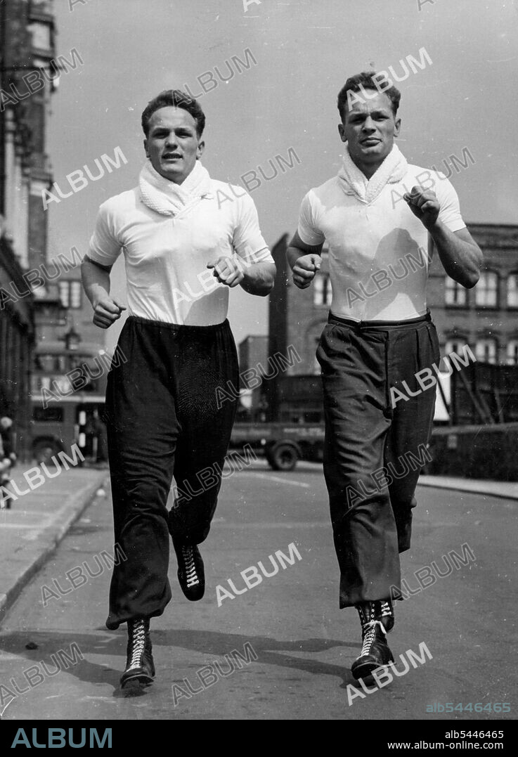 They'll Knock 'Em From The Old Kent Road -- Henry Cooper (on left) and his twin brother near their Old Kent doing a little roadwork near their Old Kent Road training quarters. Both are heavyweights. Jim Wicks, Manager of 20 year old boxer Henry Cooper, has refused an offer of $5,000 from film stars Donald Houston and Stanley Baker, for a half-share of the contract of Cooper, who comes from Bellingham, Kent. Henry is the identical twin brother of George Cooper, who is also a boxer. January 10, 1955. (Photo by Reuterphoto).