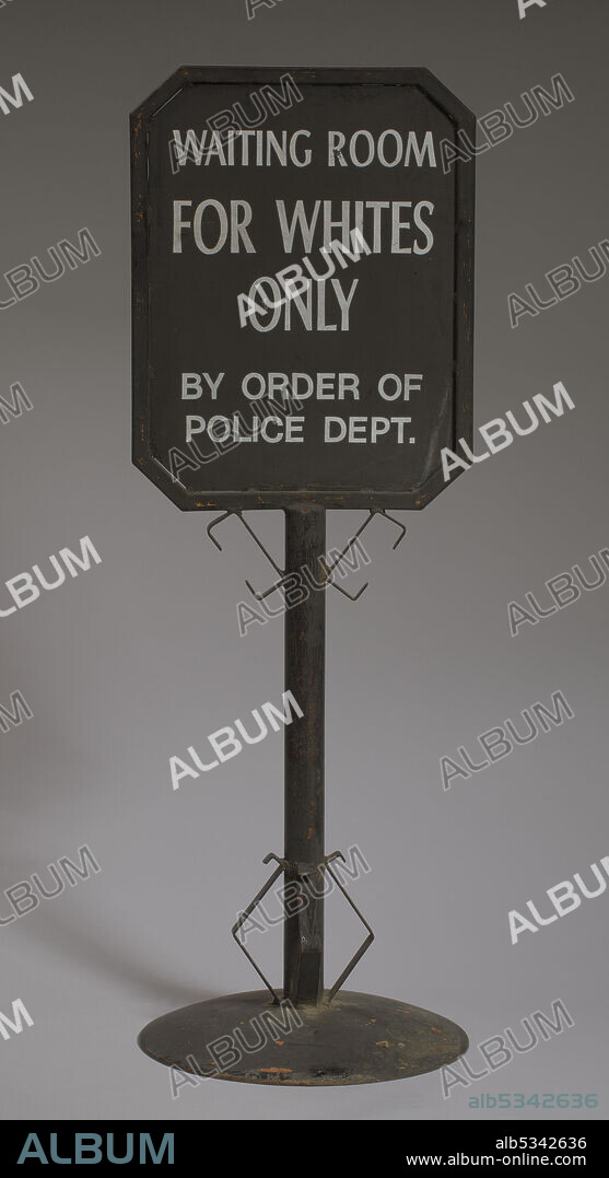Under segregation in the United States, Black people were prohibited from using the same facilities as whites. They were forced to use separate and often inferior services such as housing, medical care, education, employment, and transport. A metal double-sided black railroad sign that reads: [WAITING ROOM / FOR WHITES / ONLY / BY ORDER OF / POLICE DEPT.] in white paint. The writing is on a framed square plaque at the top of the sign. The sign post is round and has metal work at the top where the plaque meets the post and that the bottom where the post meets the base. The base is round. There is orange paint that is visible in spots where the black paint has peeled. There is also rust on the bottom of the base.