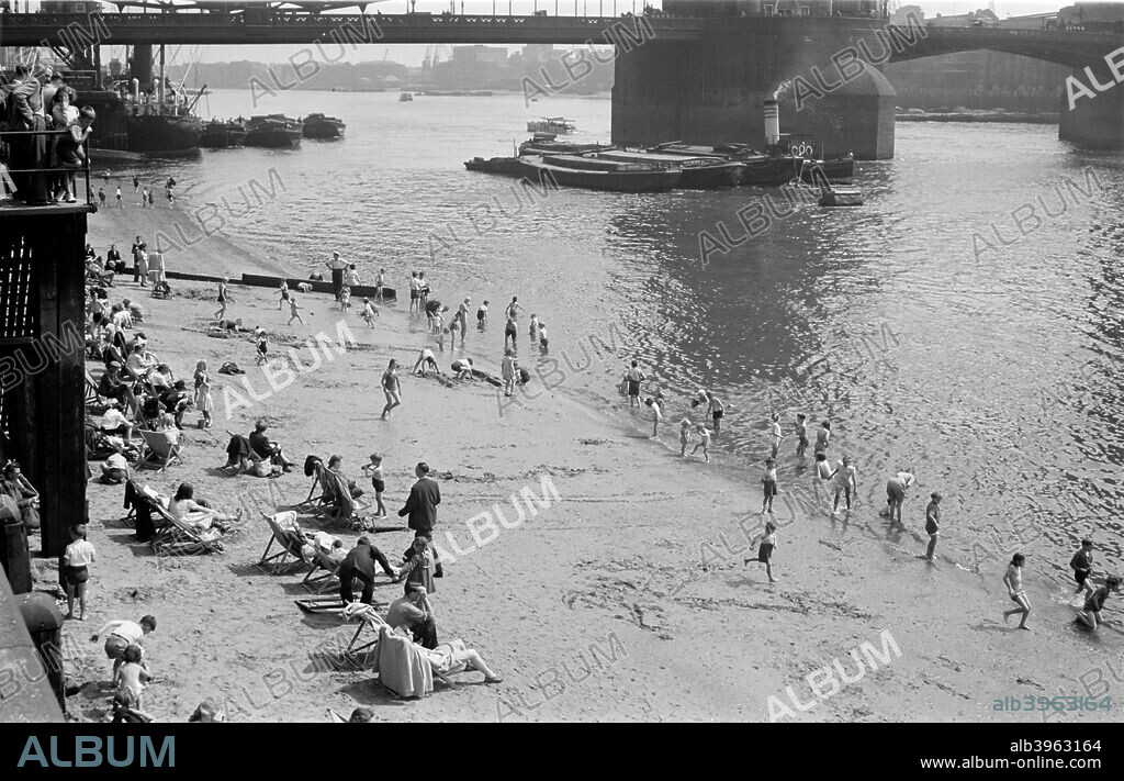 People relaxing on Tower Beach, London, c1945-c1955. Children play at the water's edge, while adults relax in deck chairs. A tug pulling barges passes the piers of Tower Bridge. The artificial beach was made just in front of the Tower of London, (now in Tower Hamlets); the sand was eventually washed away.