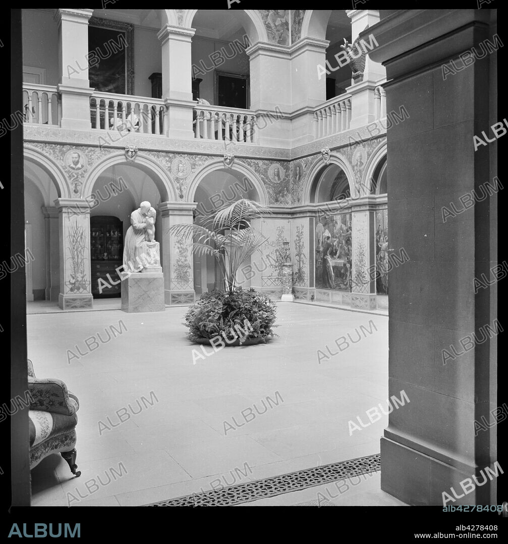 URSULA CLARK. Interior, Wallington Hall, near Cambo, Northumberland, c1955-c1980. An interior view of the central hall in Wallington Hall, showing two arcaded sides, one of them blind. The country house was built in 1688 for Sir William Blackett on the site of an earlier Fenwick house. It was remodelled for Sir William Caverley Blackett between 1735 and 1745, most likely by Daniel Garrett. Further alterations were made by John Dobson in 1853-1854. The house has a square plan around a courtyard, now a central hall roofed over in the 19th century. Arcades were added on all sides on the central hall, making the original courtyard smaller, and the north an south arcades are blind. A stair was added on the south side, behind the arcade, leading to the baclonied first floor above. The ground floor is adorned with murals by William Bell Scott, in the style of the Pre-Raphaelites. One panel was completed by John Ruskin. In the background is a marble sculpture of a woman and baby, who is stood on a plinth.