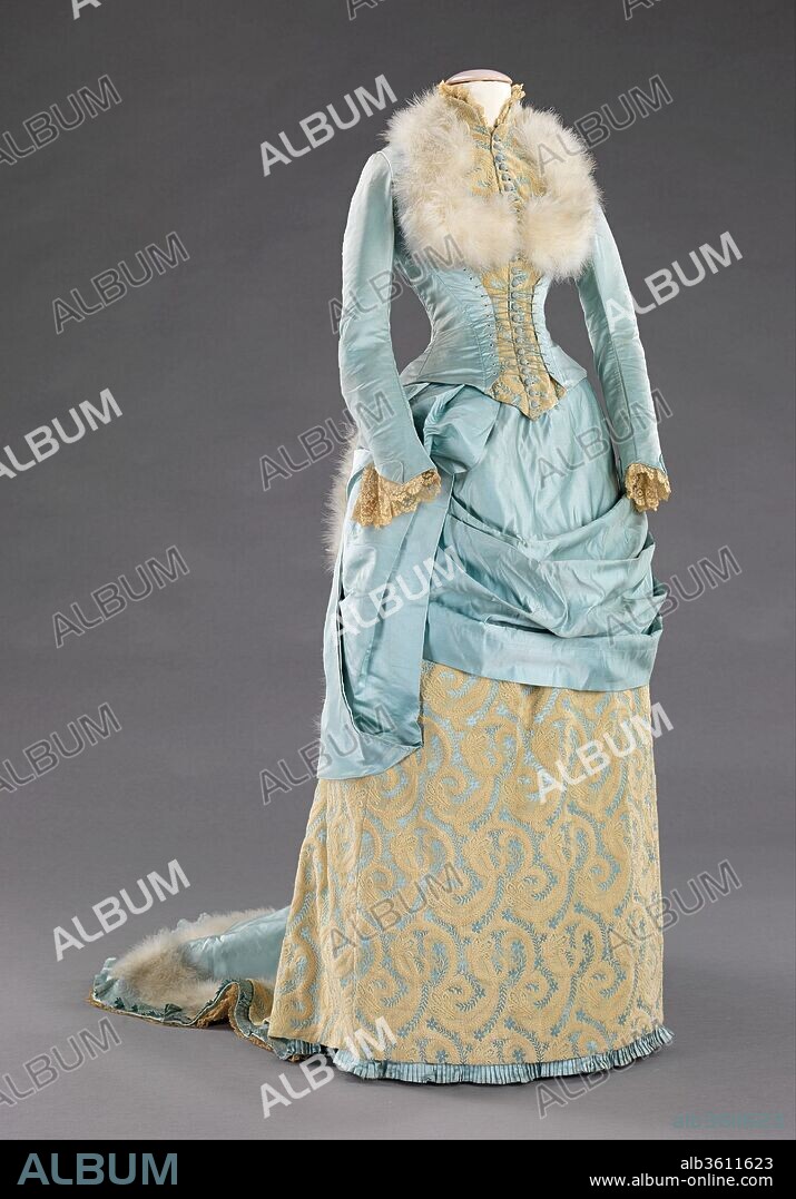 Evening dress. Culture: American. Department Store: R. H. White & Co. (American, Boston, Massachusetts). Date: 1885.
The bustle silhouette, although primarily associated with the second half of the 19th century, originated in earlier fashions as a simple bump at the back of the dress, such as with late 17th-early 18th century mantuas and late 18th- early 19th century Empire dresses.  The full-blown bustle silhouette had its first Victorian appearance in the late 1860s, which started as fullness in skirts moving to the back of the dress.  This fullness was drawn up in ties for walking that created a fashionable puff.  This trendsetting puff expanded and was then built up with supports from a variety of different things such as horsehair, metal hoops and down.  Styles of this period were often taken from historical inspiration and covered in various types of trim and lace.  Accessories were petite and allowed for the focus on the large elaborate gowns.  Around 1874, the style altered and the skirts began to hug the thighs in the front while the bustle at the back was reduced to a natural flow from the waist to the train.  This period was marked by darker colors, asymmetrical drapery, oversize accessories and elongated forms created by full-length coats.  Near the beginning of the 1880s the trends altered once again to include the bustle, this time it would reach its maximum potential with some skirts having the appearance of a full shelf at the back.  The dense textiles preferred were covered in trimming, beadwork, puffs and bows to visually elevate them further.  The feminine silhouette continued like this through 1889 before the skirts began to reduce and make way for the S-curve silhouette.
