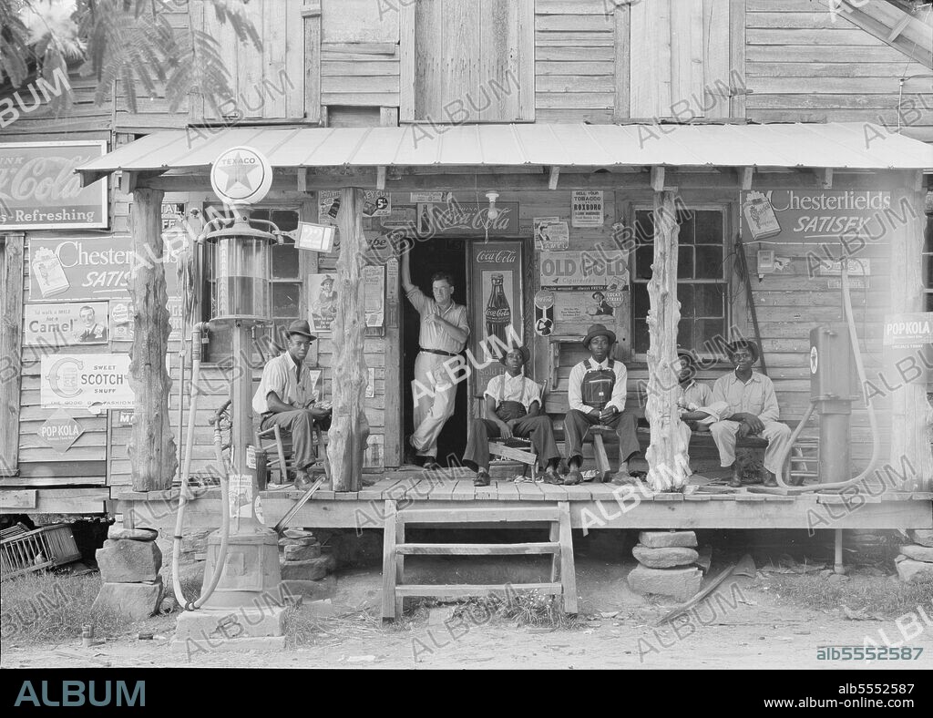DOROTHEA LANGE. Country store on dirt road. Sunday afternoon. Note the kerosene pump on the right and the gasoline pump on the left. Rough, unfinished timber posts have been used as supports for porch roof. Negro men are sitting on the porch. Brother of store owner stands in doorway. Gordonton, North Carolina.