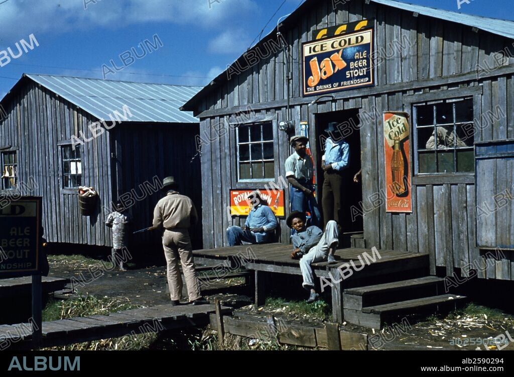 Living quarters and "juke joint" for migratory workers, sitting around during a slack season; Belle Glade, Florida, USA. 1941. Photograph shows man standing to left with gun. Signs advertise Ice Cold Jax Ale Beer Stout and Ice Cold Coca Cola.