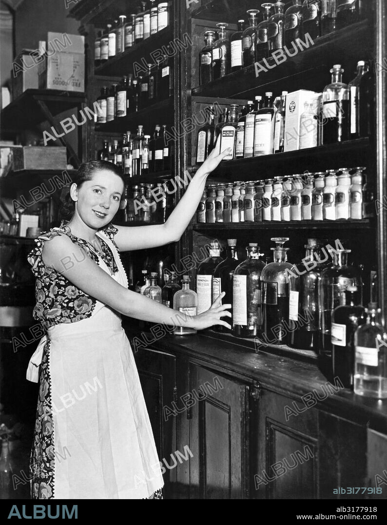 New York, New York:  1929 Marie Bard, 22, who is the model for the 1930 Red Cross poster and US postage stamp illustrated by artist Lawrence Wilbur. She doesn't smoke, doesn't stay up late, and cooks and cleans for her mother, a pharmacist, and her father, a coffee and tea importer, in Astoria, Queens. Here she is helping her mother out in the drugstore.   © Underwood Archives / The Image Works.