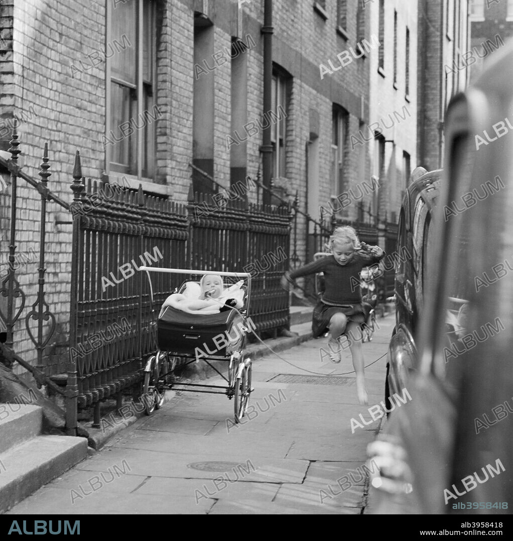 Girl skipping, London, 1960-1965. A girl jumping over a skipping rope on the pavement between a row of terraced houses and a row of parked cars, one end of the rope held by a baby in a pram.