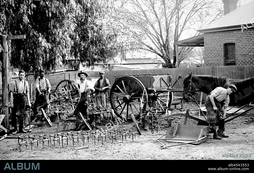 Negative - Blacksmiths' & Implement Makers' Display, Wangaratta, Victoria, circa 1915, A group of five tradsemen with a display of their products. The products shown include single and multi-furrow mouldboard ploughs, zig-zag harrows, an earth scoop, horsedrawn farm carts and tip drays, a water or feed trough and horseshoes. Four of the men are wearing leather blacksmiths' aprons and two are holding blacksmiths' hammers, while the man at the right is shoeing a horse.