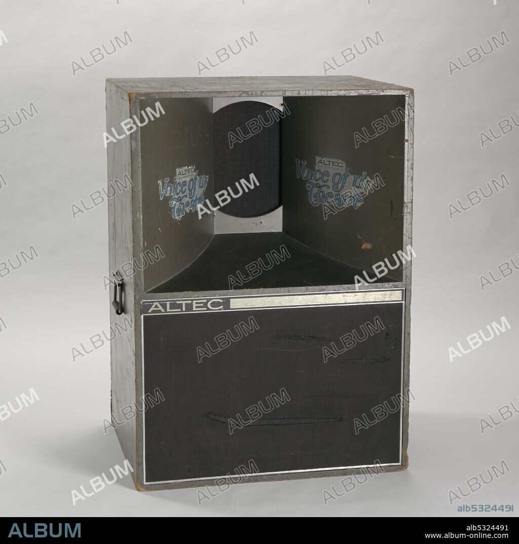 ALTEC LANSING. A speaker made by Altec. The speaker is rectangular in shape and is painted black. The top of the front of the speaker concaves inward, towards the back. The concaved sides have white and blue stickers on them that read: [ALTEC / Voice of the Theatre]. The middle of the front is black and white and reads: [ALTEC]. The bottom portion of the front is covered in mesh. The back of the speaker has outlets and the proper right and proper left sides of the speaker have handles. The bottom of the speaker has four casters at each corner.
