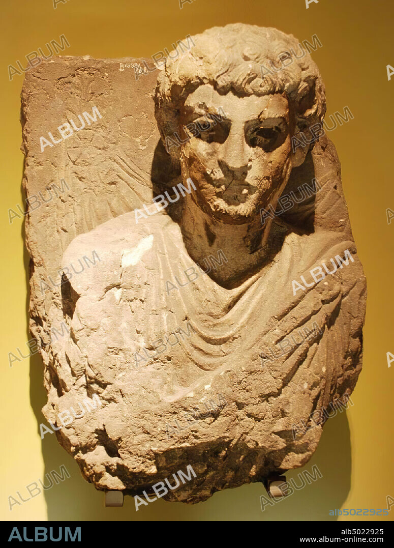 A bust of limestone. The statue represents a man with curly hair, mustache and beard, dressed in a richly folded robe. The arms have been cut away, in the background a drapery is visible. At the top right is another part of an inscription, the rest is broken away, sculpture, stone, limestone, gray, H 52 cm, W 40 cm, D 25 cm, Roman Period 200-225 AD, Syria.