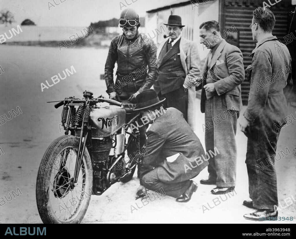 Jimmy Guthrie riding a Norton, (1930s?). A man kneels by the side of the bike inspecting it. The man dressed in a suit but without a hat is a French importer. Guthrie was a Norton works rider from 1931 to 1937, and was six times winner at the Isle of Man TT.