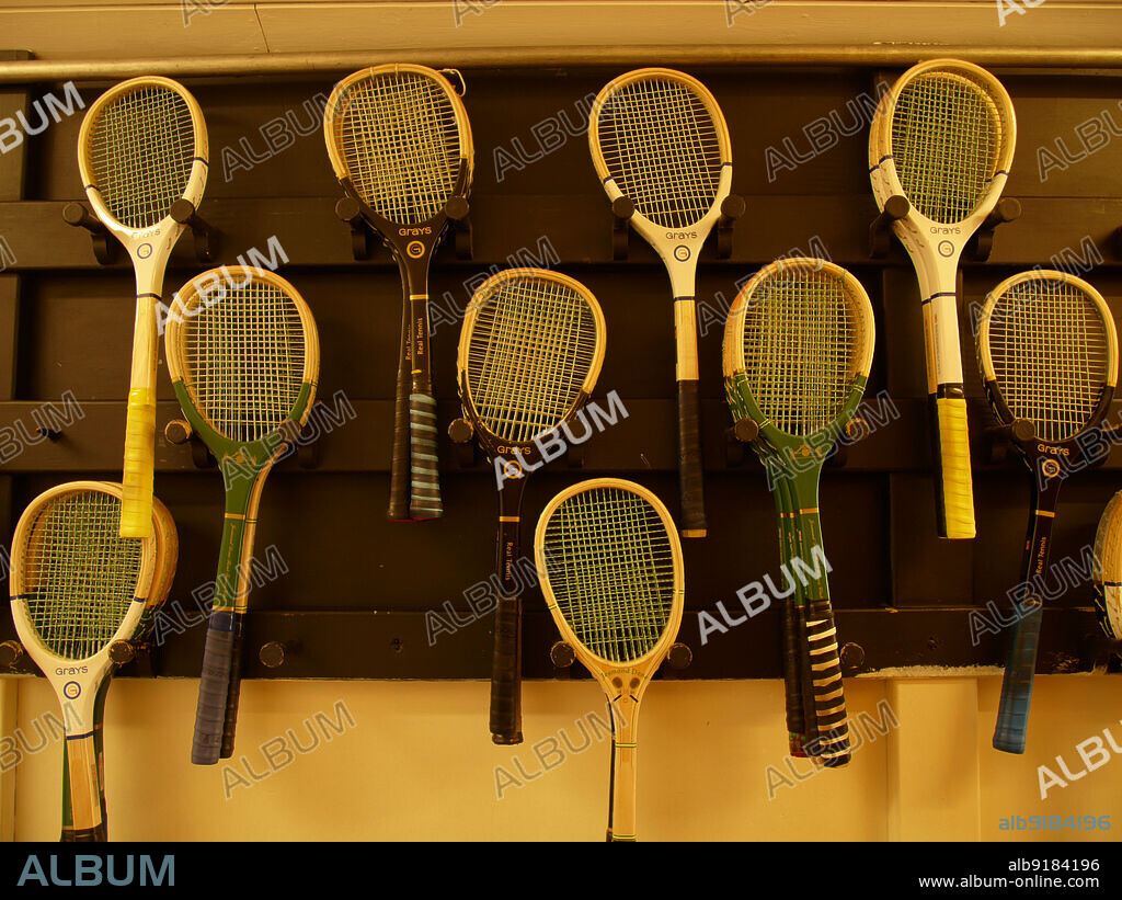 SIMON INGLIS. Jesmond Dene Real Tennis Club, Matthew Bank, Jesmond, Newcastle upon Tyne, 2010. A selection of racquets for real tennis hanging on a wall outside the court at Jesmond Dene Real Tennis Club, Newcastle upon Tyne. The tennis court was built for Sir Andrew Noble in 1894 and has been used by Jesmond Dene Real Tennis Club since its formation in 1932. Real tennis differs from the later lawn tennis in several ways, including arrangements for spectators, the construction of the tennis balls, and the style and material of the racquets. Often made from hickory or ash with tight strings, the racquets are asymmetrical and are also relatively heavy.