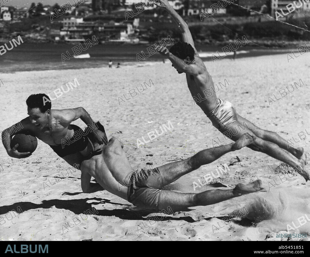 As a means of keeping warm these three members of Drummoyne Rugby Union Club played a game of football on Bondi Beach today. Johnny Denton, of Ryde, Is being tackled by John Stephenson, of Ryde, and Ian Findlay, of Filvedock. August 28, 1955. (Photo by Ronald Leslie Stewart/Fairfax Media).