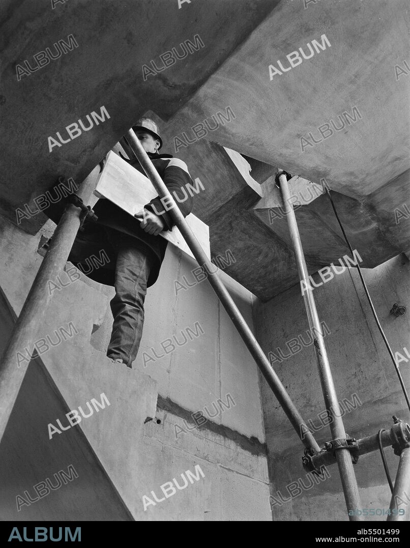 JOHN LAING PLC. Construction of a multi-storey block of 'Sectra' flats in Manchester, 09/07/1963. Looking up at a worker ascending a concrete staircase during the construction of a multi-storey block of 'Sectra' flats in Manchester. 'Sectra’ was a French prefabricated steel formwork design for flats which John Laing and Son Ltd acquired the British rights to in 1962. It was a method of using precision made steel formwork for the placing of structural concrete in 'tunnel' sections in room unit widths and ceiling heights. The units were bolted together in rows on special tracks, with the concrete poured to form the walls and floors in one operation. The formwork was internally heated to accelerate the hardening of the concrete in the mould and the sections were then lifted into position by a tower crane on the construction site.