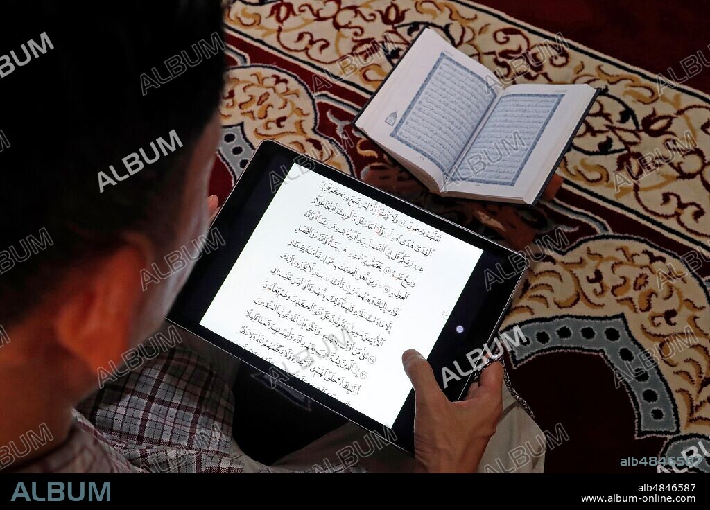 Muslim reading a digital Quran on a tablet. In the background a traditional paper Arabic Holy Quran.
