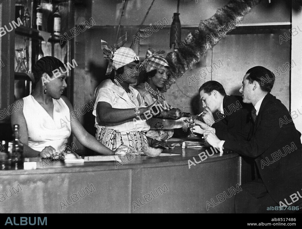 TYSKLAND BERLIN 1932-03-03. Orig. bildtext... DIE ERSTE NEGER-BAR IN BERLIN. Dem Reichshauptstädtischen Vergnügungsbetrieb hat sich jetzt eine Neger-Bar angegliedert, in der als Neuestes vom Neuen Negerinnen als Bardamen tätig sind. Das Lokal ist pariserisch und exotisch ausgestattet und wartet mit vielerlei Attraktionen auf. U.B.z. Blick auf die Bar. Foto: Keystone View Co / AB Text & Bilder / SVT / Kod: 5600 Mapp: Berlin: Caféer och restauranger -1939 G 39 J1 Två män sitter i en nyöppnad bar i Berlin och blir serverade av tre barflickor. Kontraster sites: TYSKLAND;BERLIN.