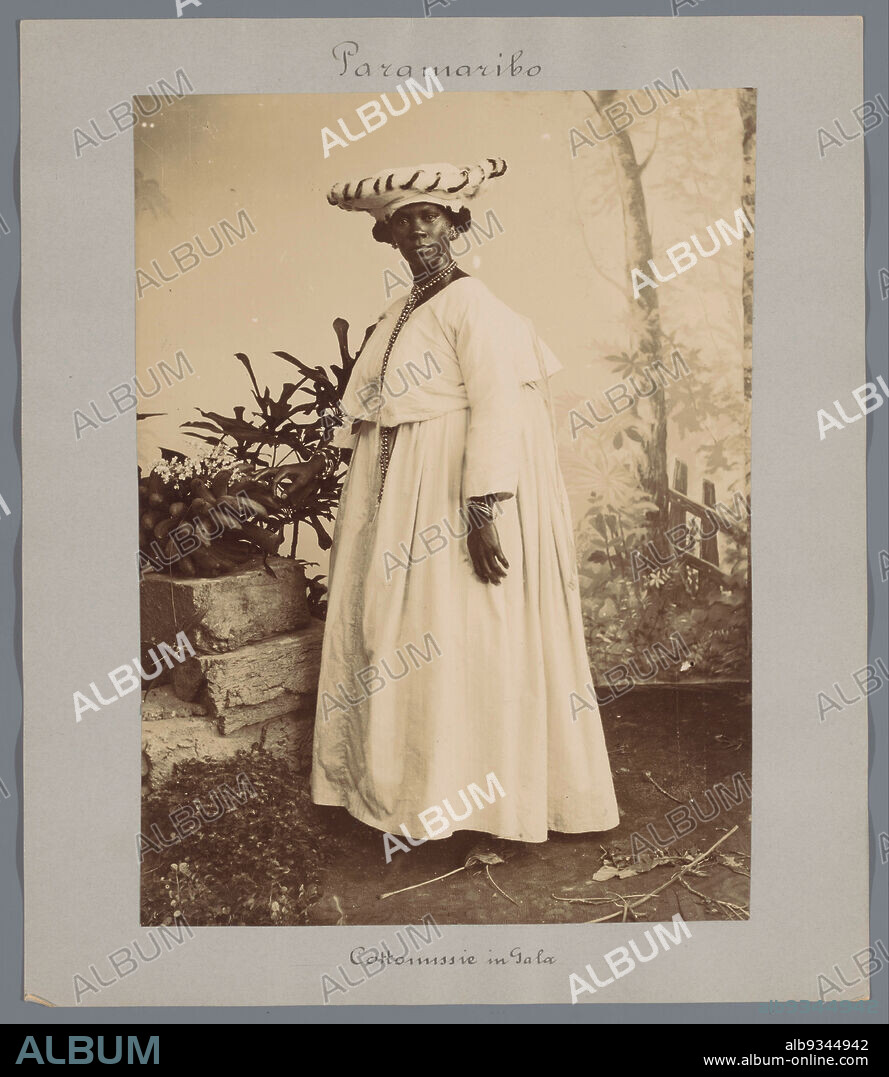 Cottomission in Gala (title on object), Portrait of a standing Surinamese woman in kotomisi. Standing near a fruit still life in front of a backdrop in the photo studio. Part of a group of 44 photographs of Paramaribo, Suriname and Curaçao, ca. 1900-1905, mounted on cardboard and inscribed., Eugen Klein, Paramaribo, 1900 - 1905, photographic support, cardboard, height 227 mm × width 166 mm.
