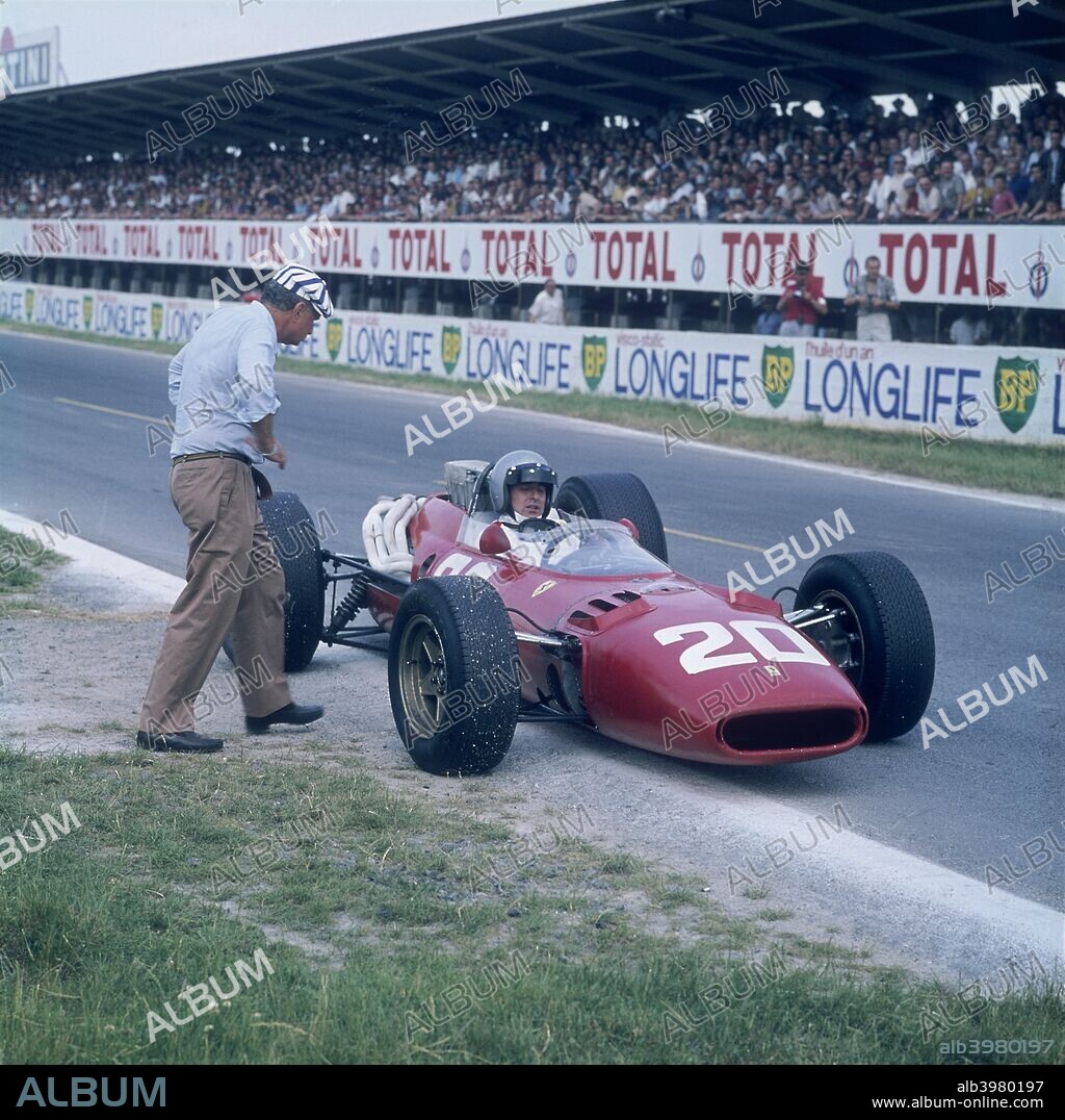 Lorenzo Bandini in a Ferrari 312, French Grand Prix, Reims, France, 1966. At the edge of the race track, with a man approaching the car. The spectator stands are behind him.