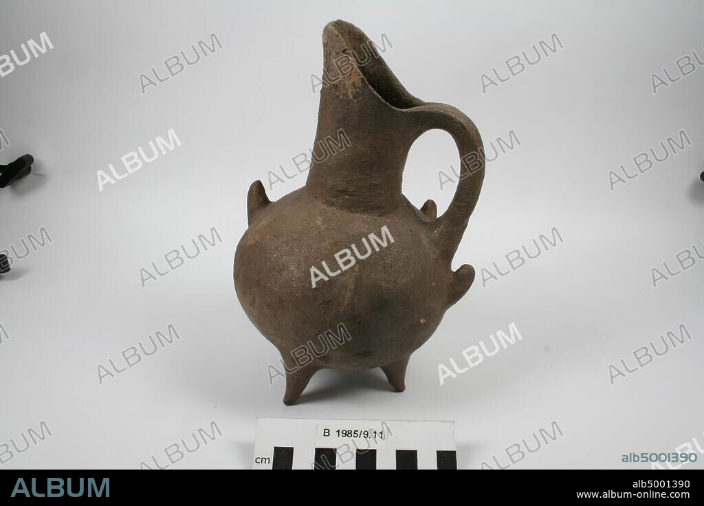 An earthenware beak cane on three legs. Four nail-shaped vertical protrusions are attached to the convex belly. The object has a wide neck with slanting edge, an ear decorated with diagonal grooves and a bronze sheen by sludge layer with mica-like magnetering., Crockery, pottery, H 21 cm, D 12.5 cm, Early Bronze Age 2800-2500 BC. , Turkey.