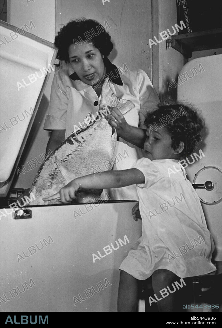This is Mrs. Ethel Robigo, daughter of Trinidad's chief justice, who will cook the meals for Princess Margaret while she is in Trinidad watching her put away huge fish in the deep-freeze is her daughter Margaret. January 31, 1955. (Photo by Daily Mirror).