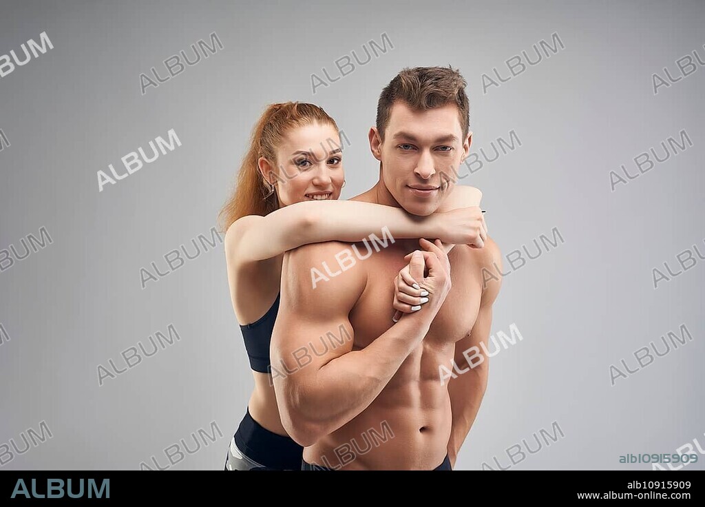Side view of muscular shirtless man and reheaded woman with ponytail in sportswear hugging man from behind against grey background.