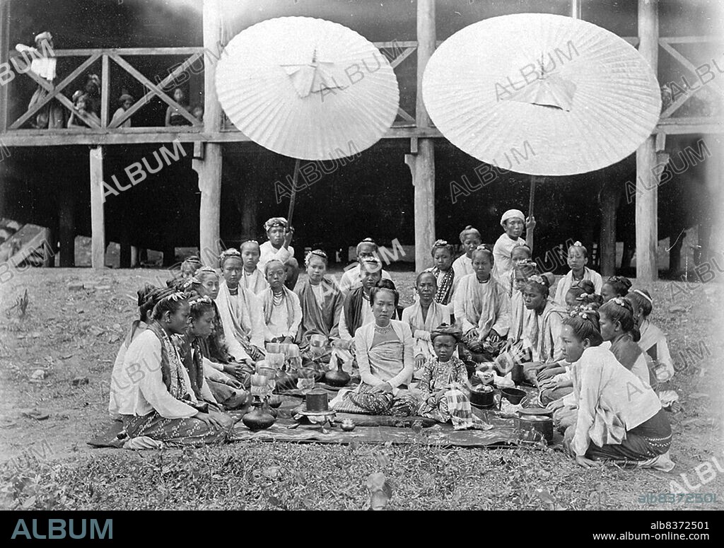 Photograph of a Sawbwas wife and child at Wuntho in Burma (Myanmar) from the Elgin Collection: 'Autumn Tour 1898', taken by Felice Beato c.1891. The portrait was taken in front of a house at Wuntho, showing the wife and child of the Wuntho Sawbwa, a Shan feudal lord, seated on the ground with food baskets. Attendants are arranged around the pair in a semi-circle and two servants hold umbrellas over the group.<br/><br/>. The woman wears a striped silk <i>hta-mein</i> (wrap-around skirt), a close-fitting jacket of fine muslin or cotton known as an <i>ein-gyi</i>, and necklaces. During the Konbaung Dynasty (1752-1885), rich jewellery, fine fabrics such as silk, and garments such as her jacket were reserved for court officials and their wives by sumptuary laws. After the fall of the Burmese monarchy they were worn by the wealthy.<br/><br/>. Wuntho or Waing Hso was a native state of Upper Burma when Burma (Myanmar), was under British control. It had an area of around 6,200 square kilometres (2,400 sq mi) with 150,000 inhabitants and lay midway between the Ayeyarwady and Chindwin rivers.