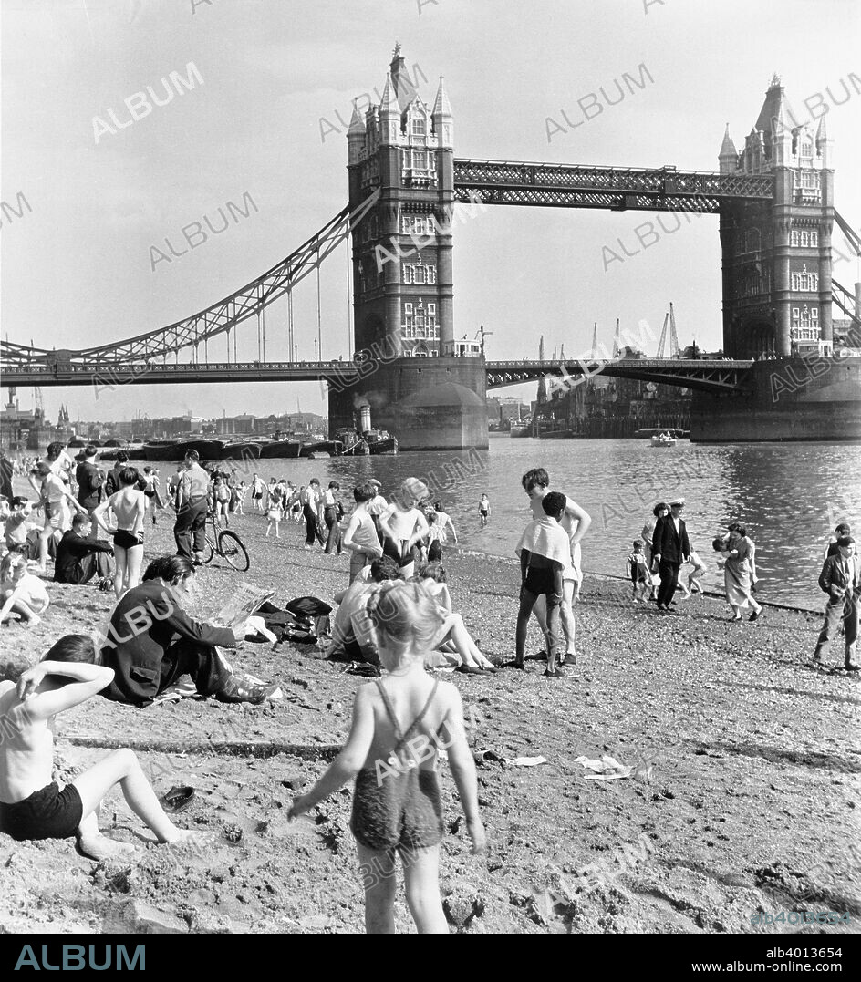 Tower Beach, London, 1952. People relaxing on an artificial beach on the River Thames, with a tug pulling barges under Tower Bridge in the background. The bridge was designed by Horace Jones.