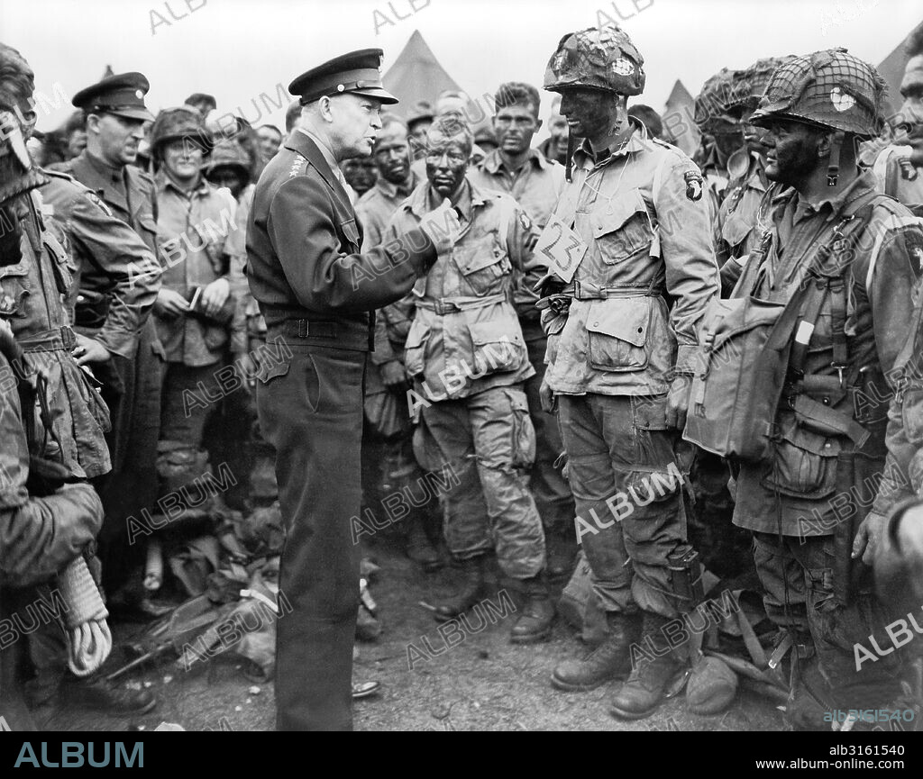 Berkshire, England: June 5, 1944 General Dwight D. Eisenhower talking with American paratroopers on the evening of June 5, 1944, as they prepared for the Invasion of Normandy. The men are part of Company E, 502nd Parachute Infantry Regiment, at the 101st Airborne Division's camp in Greenham Common. Photo includes Sgt. Russell Wilmarth, behind Eisenhower's chin, Lt. Wallace C. Strobel with a '23' tag, Ralph 'Bud' Thomas, to the left of Strobel, (and probably Corporal Donald E. Kruger, in front row, far right, wearing a musette bag on his chest.