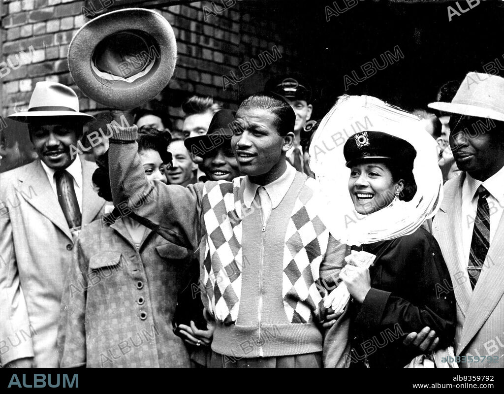 "Sugar" Ray Robinson Arrives - Showgirl wife Edna Mac. With his entourage and smiling wife, world middleweight boxing champion "Sugar" Ray Robinson made a gay arrival in London after his string of continental victories. Wearing a colorful diamond - checked cardigan, "Sugar" waved a greeting to the crowd. He will defend his title against Randolph Turpin tomorrow night. June 4, 1951.
