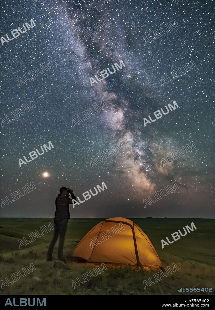 A person poses for a scene in Grasslands National Park, Saskatchewan, Canada, of stargazing with binoculars under the Milky Way on a dark moonless night. Grasslands is perfect for stargazing as it is a Dark Sky Preserve and the horizon is vast and unobstructed. Mars is bright to the left and the galactic centre is to the south at right. The view is overlooking the Frenchman River Valley.