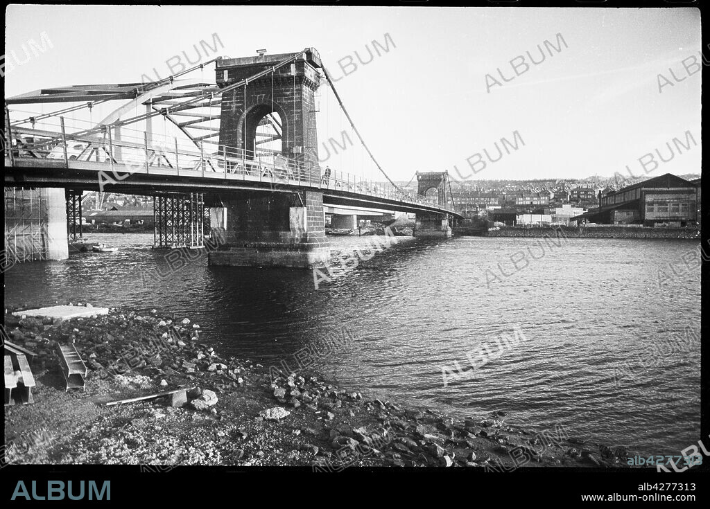 URSULA CLARK. Old Scotswood Bridge, Gateshead, Tyne & Wear, c1955-c1967. Known locally as the Chain Bridge, the Old Scotswood Bridge was a suspension bridge with two large stone river piers, designed by John Green and opened in April 1831. When completed it was the first bridge over the River Tyne on Tyneside to be built in the industrial era. The bridge was demolished in 1967 and replaced by a new bridge, seen under construction in the background.