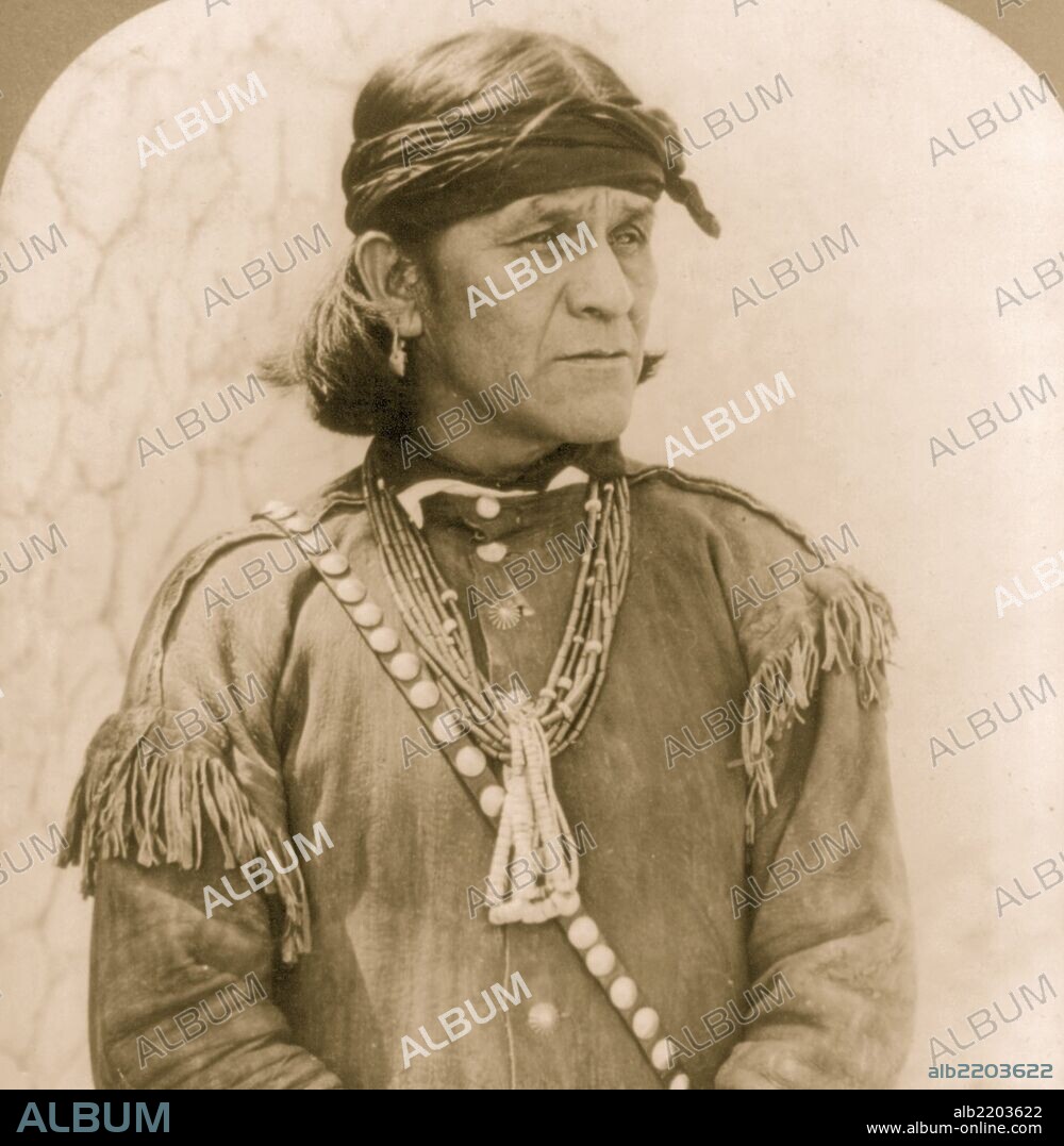 Male Hopi Indian, wearing a head band, an earring, necklaces and probably a buckskin jacket. Half length, standing, facing right.