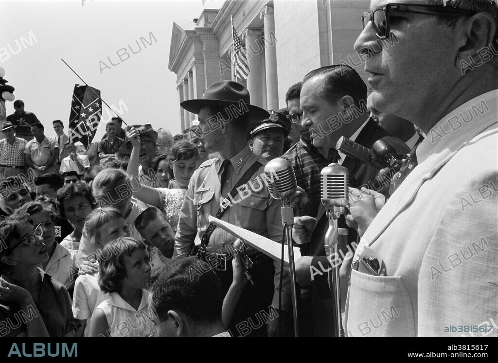 Entitled: "Little Rock, 1959. Rally at state capitol" shows a group of people, one holding a Confederate flag, surrounding speakers and National Guard, protesting the admission of the "Little Rock Nine" to Central High School. The Little Rock Nine were a group of nine African-American students enrolled in Little Rock Central High School in 1957. Their enrollment was followed by the Little Rock Crisis, in which the students were initially prevented from entering the racially segregated school by Orval Faubus, the Governor of Arkansas. After the 13th Amendment abolished slavery in America, racial discrimination became regulated by the so-called Jim Crow laws, which mandated strict segregation of the races. This legislation that mandated segregation lasted to the mid-1960s. Institutionalized racial segregation was ended as an official practice by the efforts of such civil rights activists as Clarence Mitchell, Rosa Parks, and Martin Luther King Jr., working during the period from the end of WWII through the passage of the Voting Rights Act and the Civil Rights Act of 1964. Photographed by John T. Bledsoe, August 20, 1959.