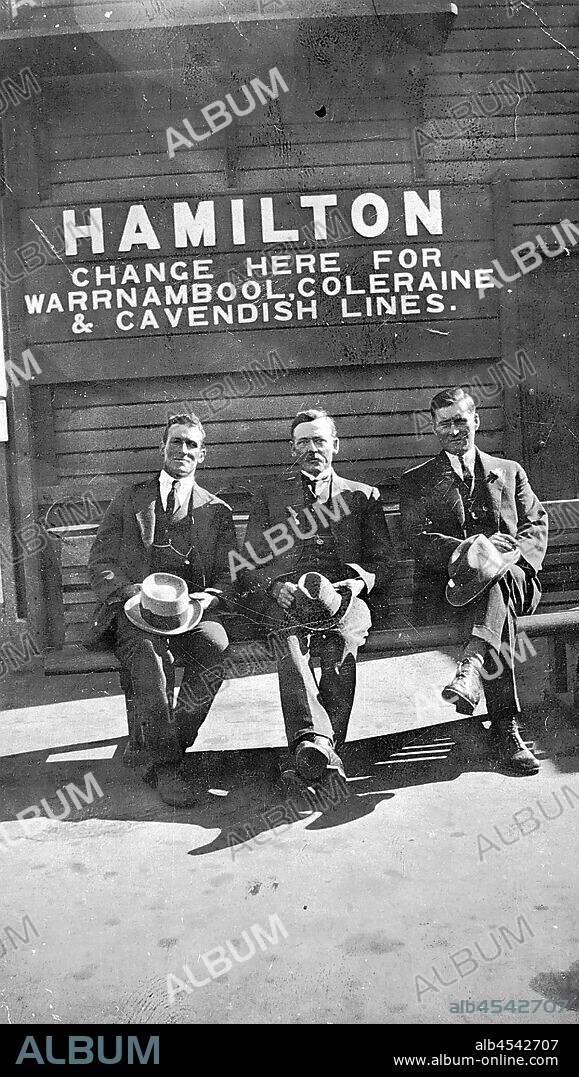 Negative - Hamilton, Victoria, circa 1920, Three men sitting on a bench at Hamiliton Railway Station. That are sitting below a sign that says 'Hamilton - Change here for Warrnambool, Coleraine & Cavendish lines'.