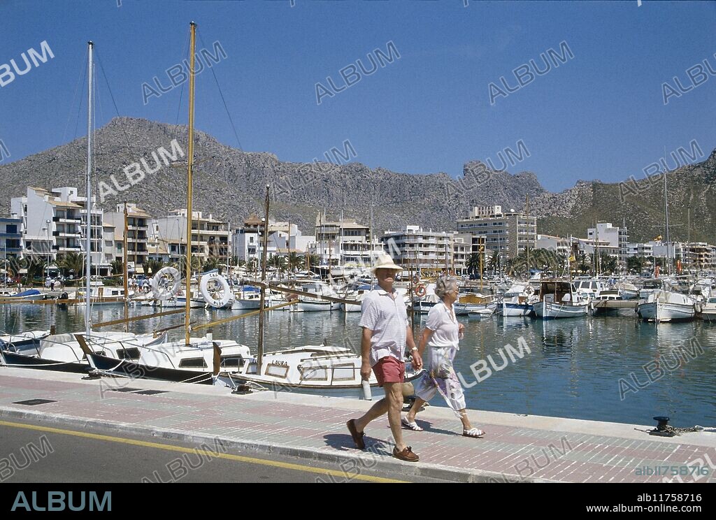 A couple striding along one of the pontoons between the many boats tied up in the marina of Port de Pollenca *** Local Caption ***.