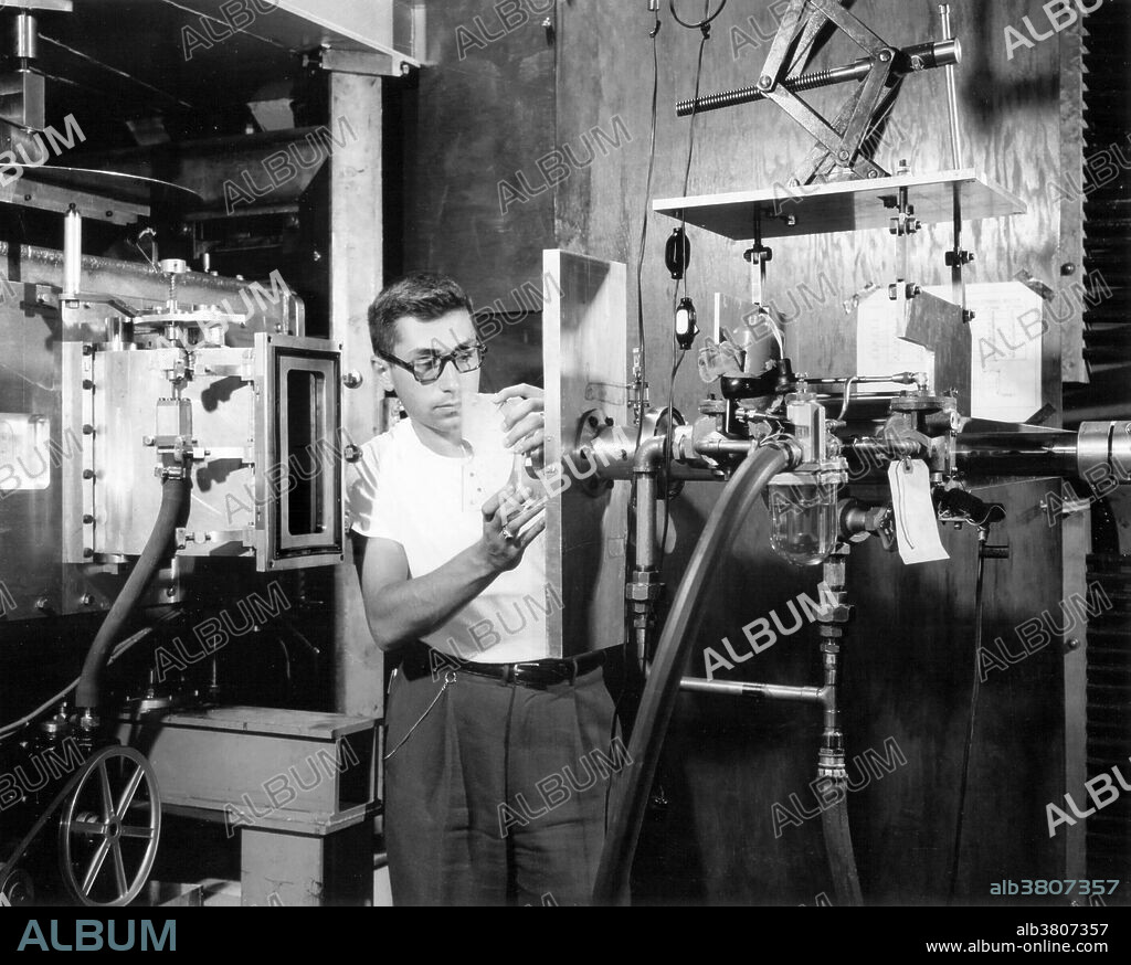 1954. A Brookhaven Lab physicist readies a piece of plastic which will be bombarded by billion-volt protons in the Cosmotron at Brookhaven National Laboratory. When the rectangular slab is moved tightly against the rectangular groove, air is pumped out of the small chamber. An operator in the control room can then set up automatic equipment which, after the Cosmotron is started up, will push the target into the path of the protons as they circulate in the vacuum chamber (left). Fragments of the nuclei target atoms are trapped in research equipment for studies of the structure and properties of the atomic nucleus. The Cosmotron ceased to operate December 31, 1966.