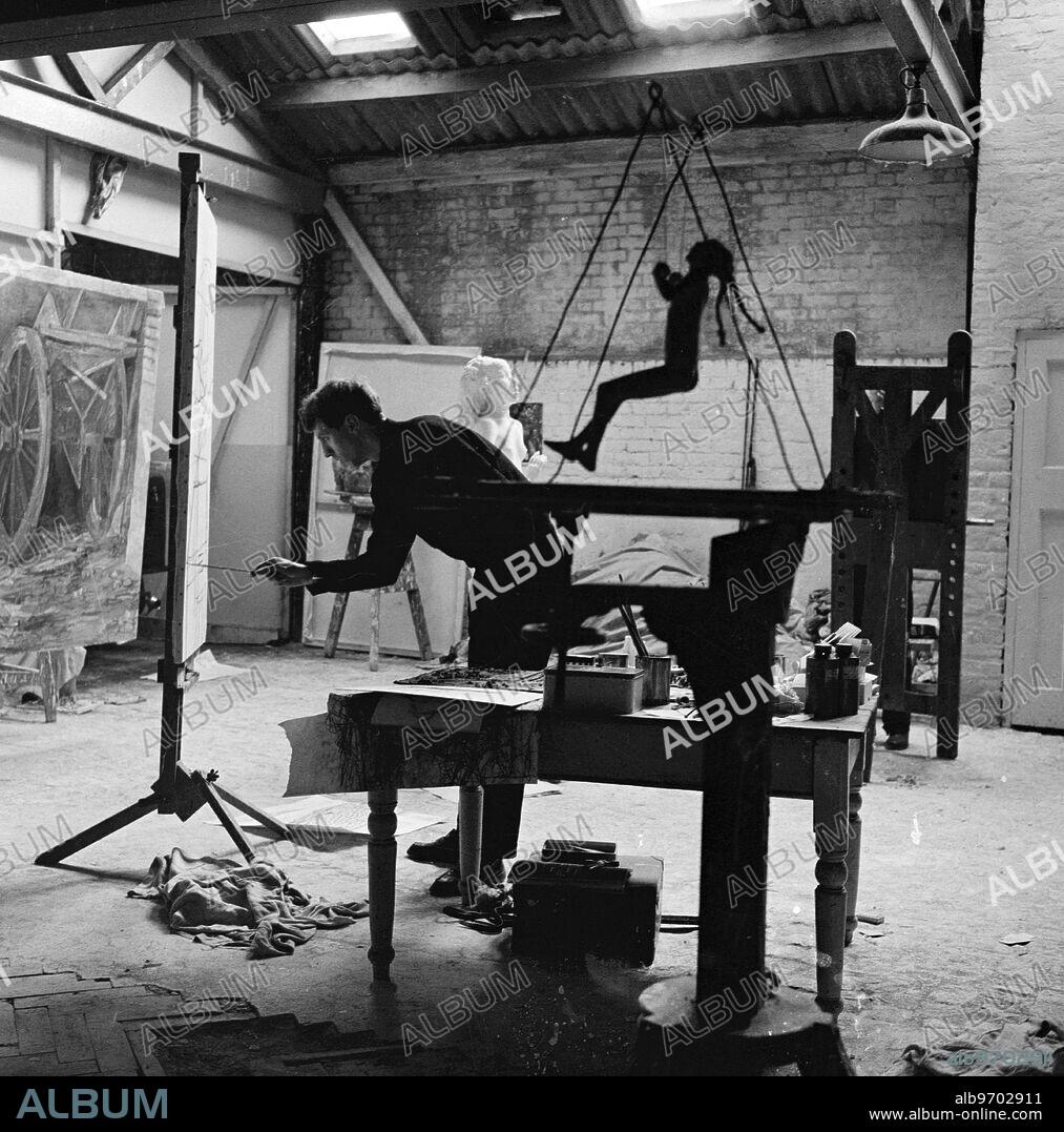 Original 1950s caption. Art in the garage . by Frank Monaco. Derrick Greaves , 27 , is a painter , George Fullard , 31 is a sculptor . Both come from Sheffield , in the North of England . Both , after winning scholarship to Italy and wishing they could have stayed there , have ended by living in London , centre of the English arts , and pooling their sometimes slender resources to hire a large studio behind a garage off the Fulham Road . They are both busy preparing for one-man exhibitions ( ' if ' , as George puts it , we can find one man reckless enough to let us have one ' ) . This is a job needing a long- term patience , and subject to fits of self - criticism and depression , which the average citizen happily does not have to possess or endure . PICTURE SHOWS: With Fullard's Girl On Swing in the foreground , Greaves blocks in new painting ... November 1954. Copy of original interview with both artists is held on file at TopFoto.