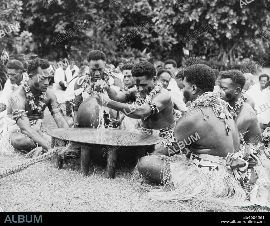 Fiji, Performing a 'sevusevu' ceremonyFive Fijian men sit in a circle around a wooden 'tanoa' as they participate in a traditional 'sevusevu' welcoming ceremony. Naked from the waist up, the men wear face paint, grass skirts and leis made from leaves. The central figure strains a mixture of water and powdered kava root (Piper methysticum) through a cloth to remove any grit, before distributing the infusion. Caption reads: The Yanqona ceremonyThis is a greatly revered ceremony of welcome though it is in no sense religious. Yanqona is also the traditional drink of the Fijian people. It is made from the root of a plant which is grated when fresh and mixed with water. The ceremony is long and solemn. Yanqona is liked by some Europeans, but to others it tastes like dish water mixed with a generous amount of Gregory powder, 1965. 2005/010/1/14/46.