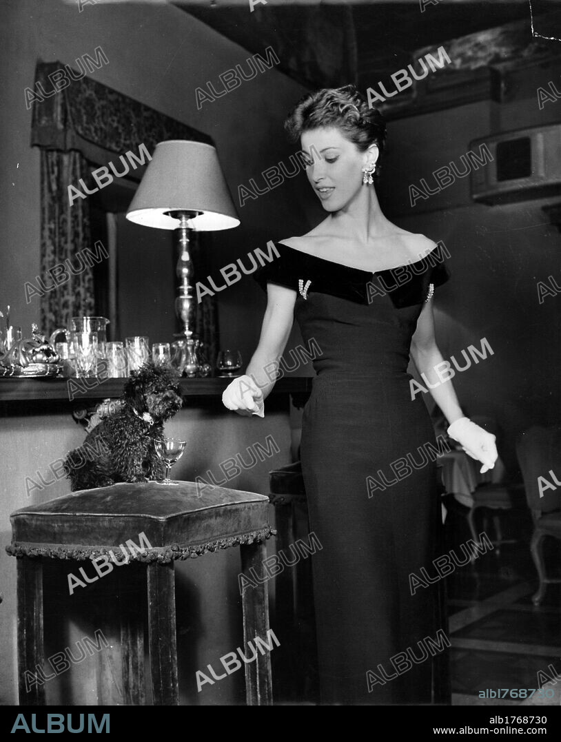 Mannequin posing. A woman with a low-cut dress and gloves looking at a poodle dog. 1955.