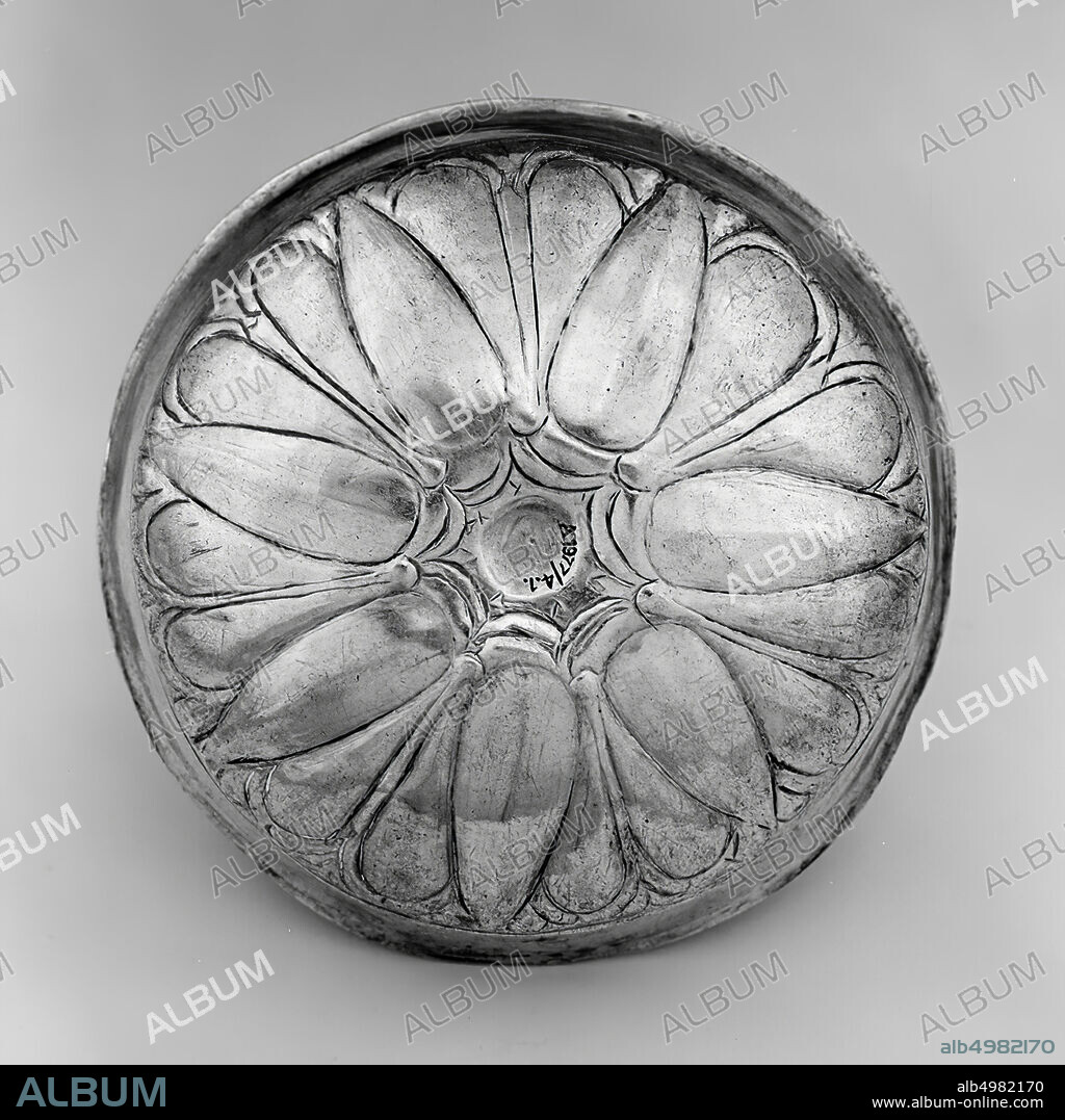 A small silver bowl with an S-shaped wall. The bowl has an omphallos bottom and a very thin regular wall. The bottom is decorated with a rosette, accented with engraved lines. The rosette has seven complete leaves that run in a point. Two rounded leaves can be seen between each leaf. Probably it is a representation of a lotus flower. The decoration can be seen on the inside and the outside., Crockery, metal, silver, H 4.1 cm, D 14 cm, Achemenid Period 500-331 BC, Iran.