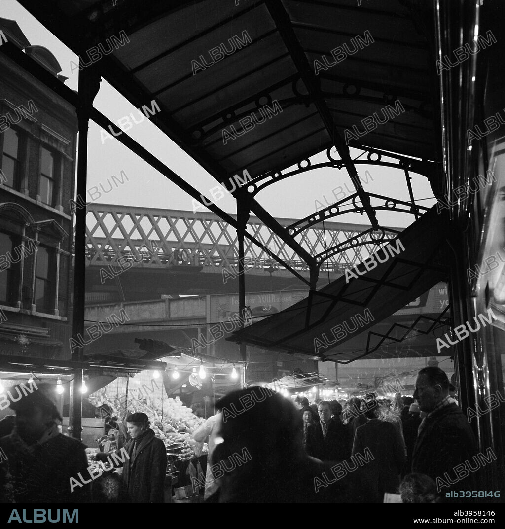 Street market in Electric Avenue, Brixton, London, 1962-1964. People shopping at twilight, seen from under one of the ornate ironwork canopies and looking towards the railway bridges crossing Atlantic Road. Electric Avenue was the first shopping area in London to be lit with electric light. It also gave its name to a song by Eddy Grant.