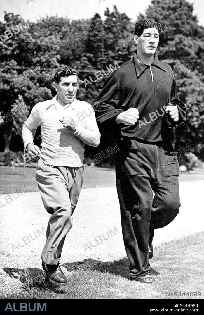 South African boxer Ewart Potgieter towers over his trainer John Holt in an exercise run at Brighton, England. Potgieter is 7ft 2in high and weighs 22½st. August 27, 1955.